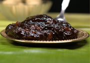 A plate of Karuppatti Halwa, a type of snack cake with a sticky and textured surface, placed on a green surface with a spoon beside it.